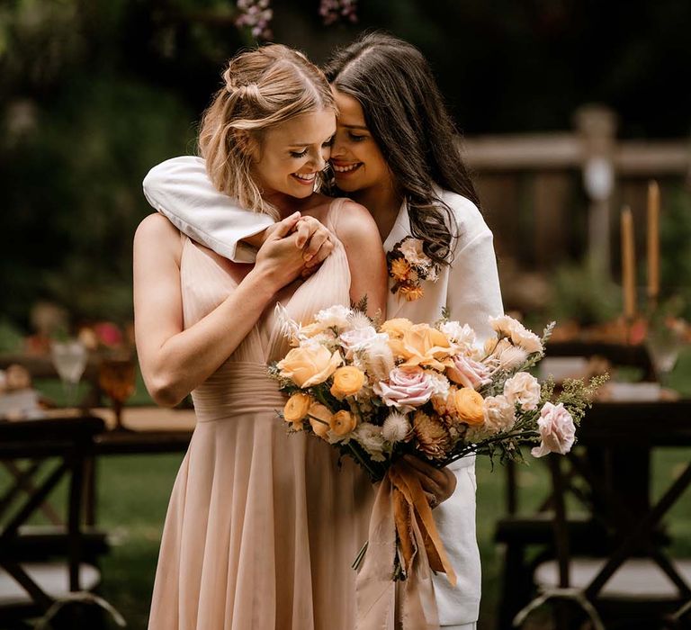 Bride in a white suit embracing her bride in a blush pink wedding dress as she holds a peeled back rose wedding bouquet 