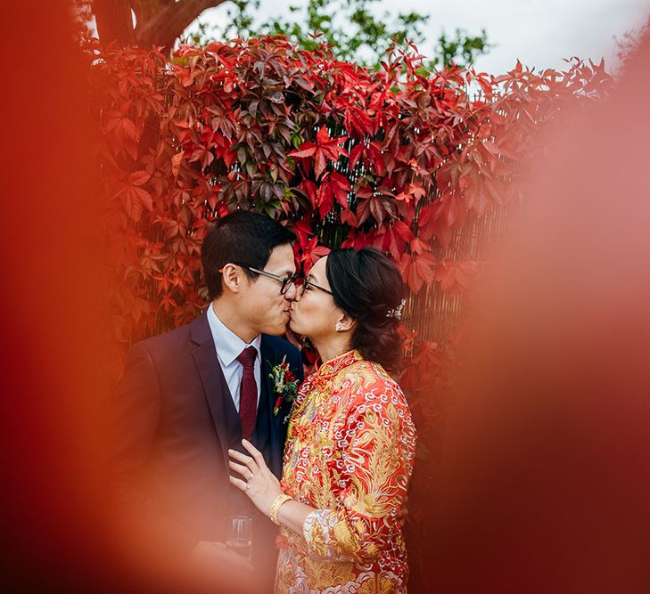 A Chinese couple in traditional Chinese tea ceremony dress kiss in front of a wall of red leaves. 