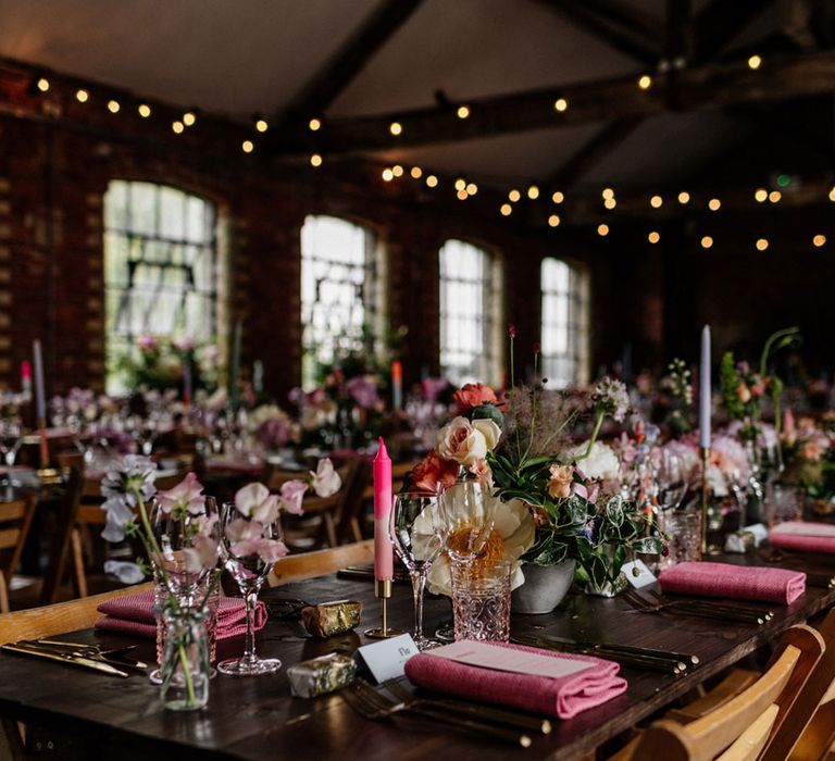 Long wooden table with folding wooden chairs, multicoloured candles, mixed florals and pink napkins in industrial room with festoon lighting for wedding breakfast at Loft Studios London