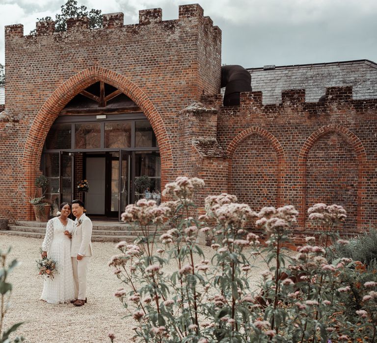 Bride in long mesh sleeve spotted Vagabond wedding dress holding mixed bridal bouquet stands next to groom in double breasted linen suit standing outside brick exterior entrance to Wasing Park