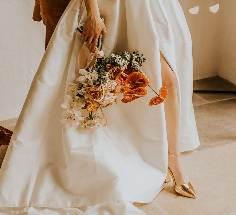 Bride in a taffeta wedding dress with front split and metallic shoes holding her orange and yellow wedding bouquet by her side 