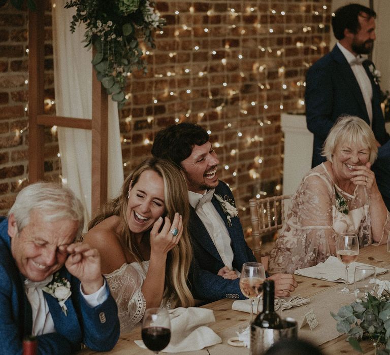 Bride in Grace Loves Lace bardot wedding dress and groom in blue jacket and bow tie laugh with wedding guests as they sit at top table in front of string fairy lights at Isle of Wight wedding with macrame wedding decor