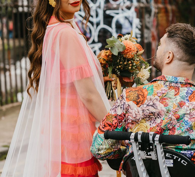 Beautiful bride in an embroidered cape veil by Rebecca Anne Designs and coral wedding dress roaming the streets of London with her disabled groom in a wheelchair 