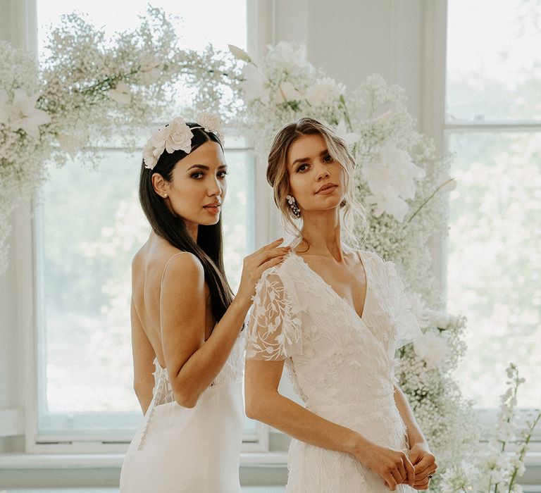 Two brides in Constellation Ame wedding dresses with lace and overlay and inserts standing in front of a white gypsophila flower arch