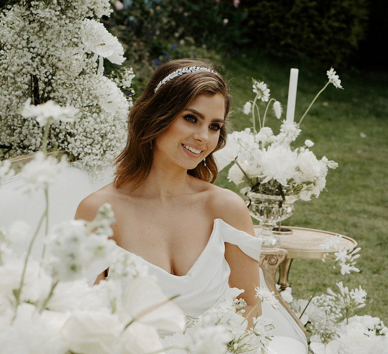 Bride with shoulder length brunette hair wearing a jewel headband in a satin structured off the shoulder wedding dress surrounded by white wedding flowers 