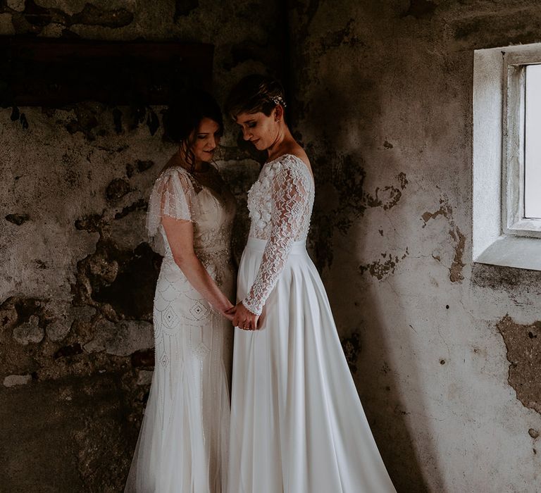 Intimate wedding photography of two brides holding hands in their vintage embellished wedding dress and appliqué gown with long sleeves and sateen skirt 