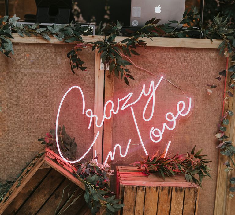 Pink neon Crazy in Love sign behind wooden crates with foliage in front of DJ booth for summer wedding at Primrose Hill Farm