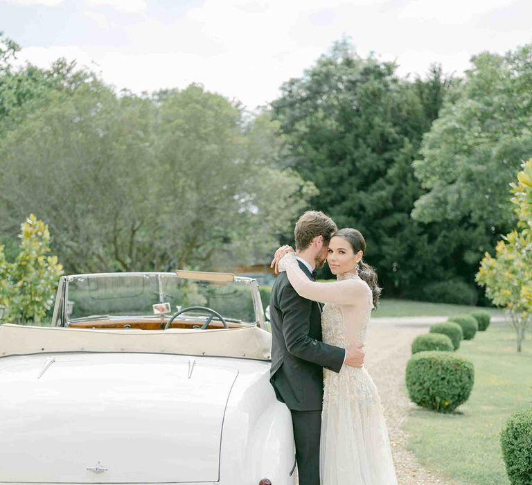 Bride in a romantic lace wedding dress with long sleeves embracing her husband in a tuxedo by their vintage wedding car 