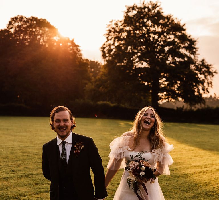 Bride in whit Halfpenny London Mayfair dress with ruffle sleeves holding mixed bridal bouquet with burgundy ribbons walks through garden holding hands with groom in three piece suit and buttonhole during holden hour at wedding in Cornwall