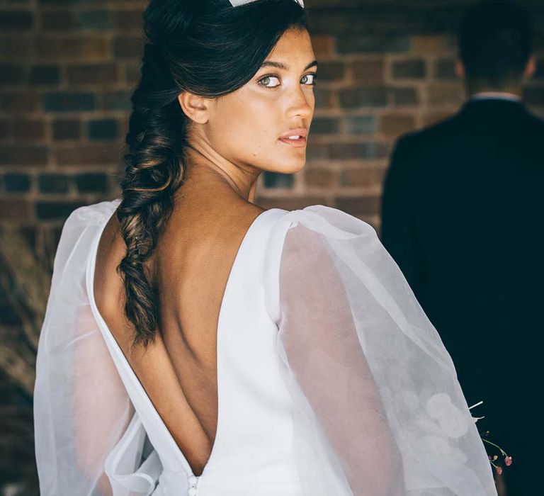 Bride in a low back wedding dress with buttons and sheer long sleeves looking over her shoulder with a plait up do and flower headband 