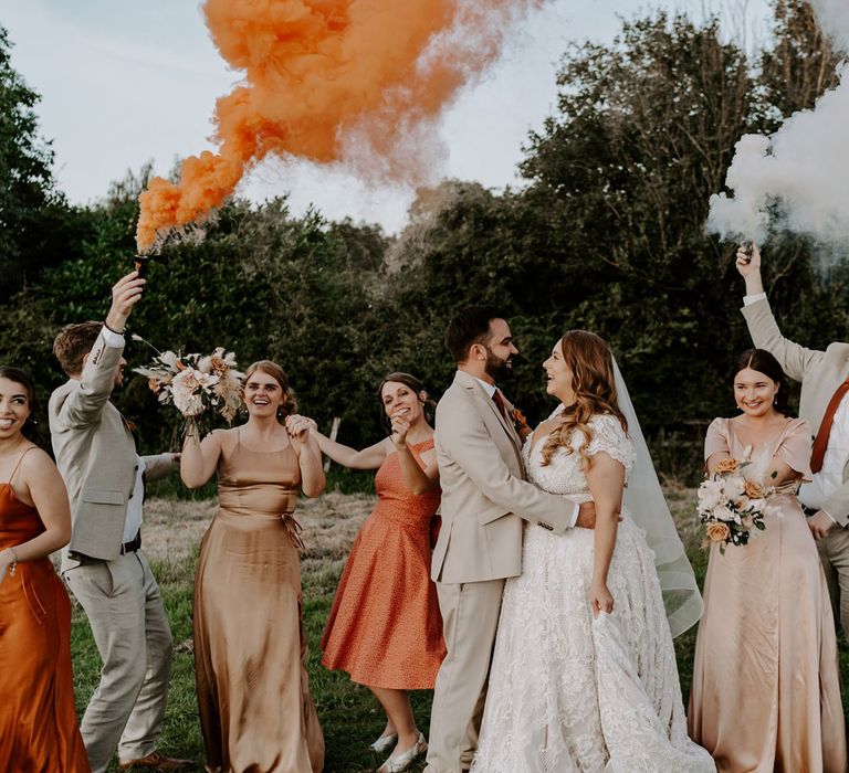 Bride in homemade lace wedding dress holds hands with groom in beige three piece suit as they stand with groomsmen and bridesmaids in field whilst holding orange and white smoke bombs