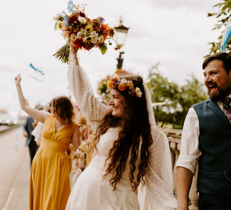 Bride lifts her colourful floral bouquet in the air as she walks down the street with her groom and wedding party