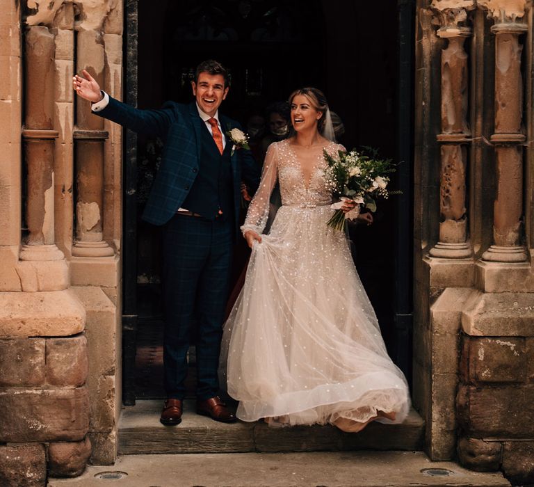 Groom in blue checked suit throws out his arms as he leaves church with bride in mesh long sleeve Julie Vino wedding dress and veil after wedding ceremony