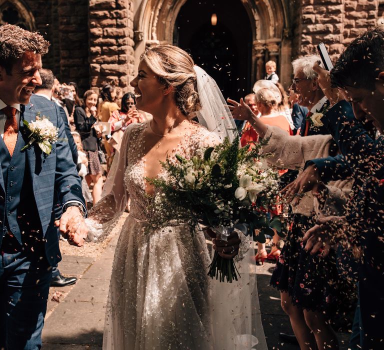 Groom in blue three piece checked suit smiles as he holds hands with bride in long sleeve embroidered Julie Vino wedding dress and veil whilst guests throw confetti