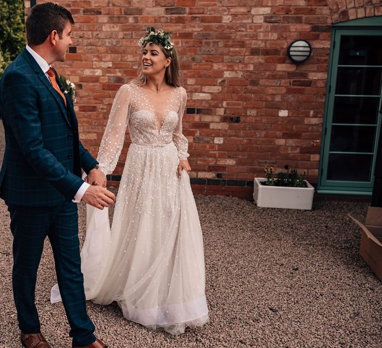 Bride in long mesh sleeve Julie Vino wedding dress and flower crown walks across drive with groom in blue checked three piece suit and floral button hole