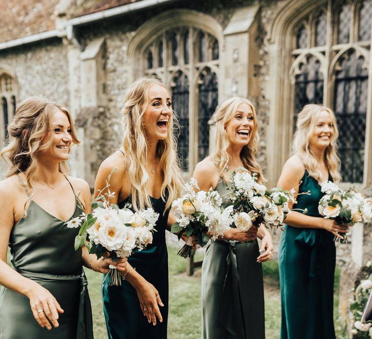 Four bridesmaids in green satin bridesmaid dresses holding white and green wedding bouquets smiles at bride stood outside of church