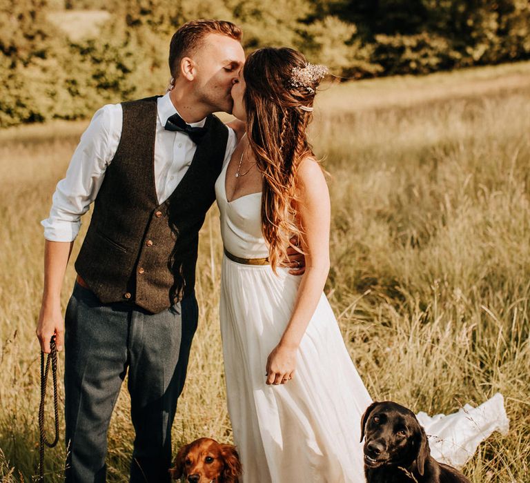 Bride and groom standing in a field kissing with their pet dogs beside them