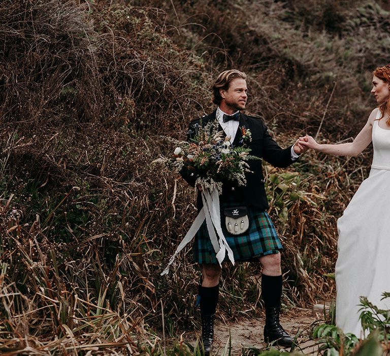 Groom in a blue and green tartan kilt holding his brides hand in a April Ross wedding dress and her boho wedding bouquet 