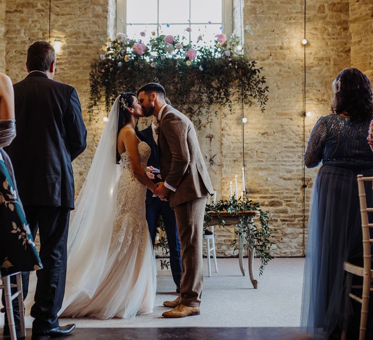 Bride in lace and tulle wedding dress with train and veil kisses groom in brown check tweed suit during stone barn wedding ceremony in the Cotswolds