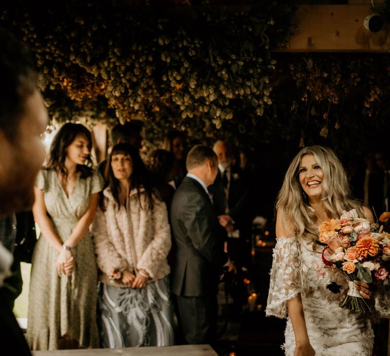 Bride in boho wedding dress holding orange, pink and white rustic mixed bouquet smiles as she walks towards groom during rustic wedding ceremony at Lyde Court