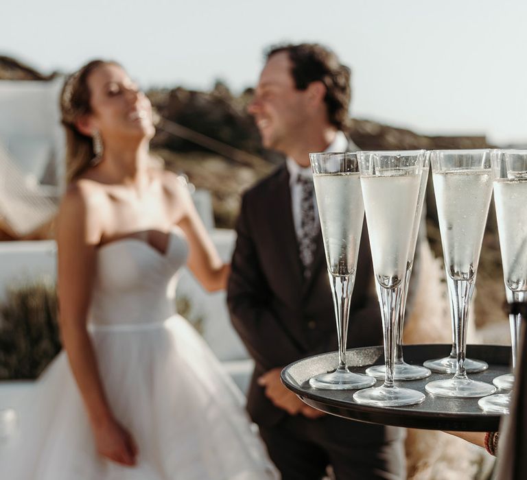 Bride & groom stand outdoors on their wedding day as a tray of champagne can be seen in front of them