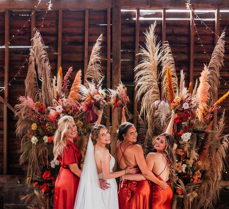 Bride in corset back wedding dress and veil stands looking over her shoulder with bridesmaids in mixed orange satin bridesmaid dresses by rustic floral installation at barn wedding ceremony all holding up dried floral bouquets 