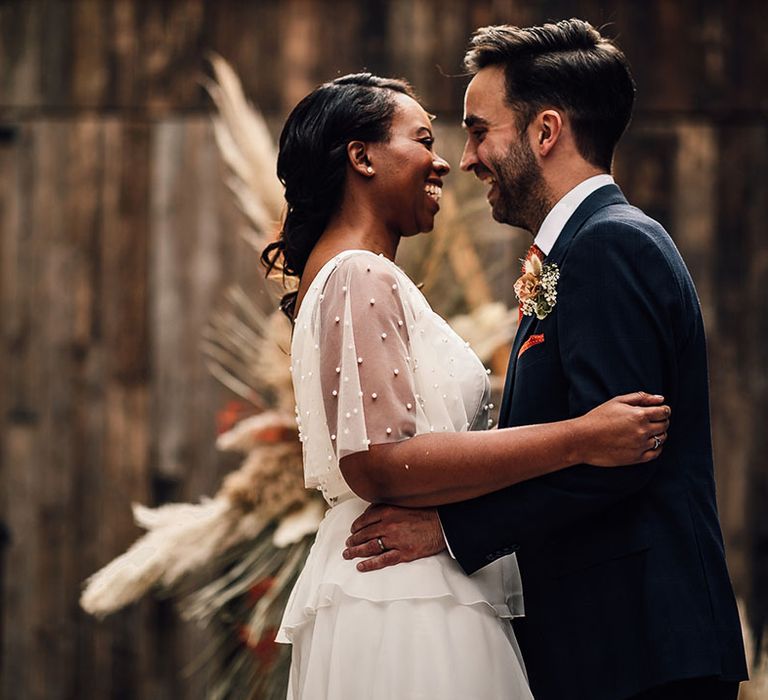 Groom in a navy suit embracing his bride in a sheer sleeve wedding dress at their 100 Barrington wedding ceremony 