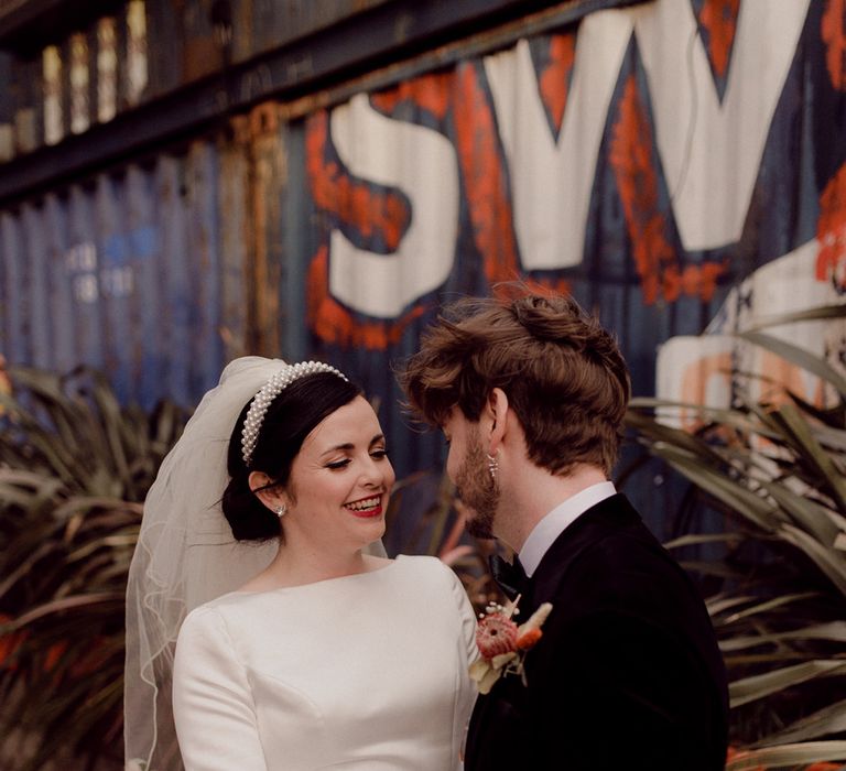 Bride in pearl headband and high neck long sleeve wedding dress stands with groom in black tie in front of colourful backdrop