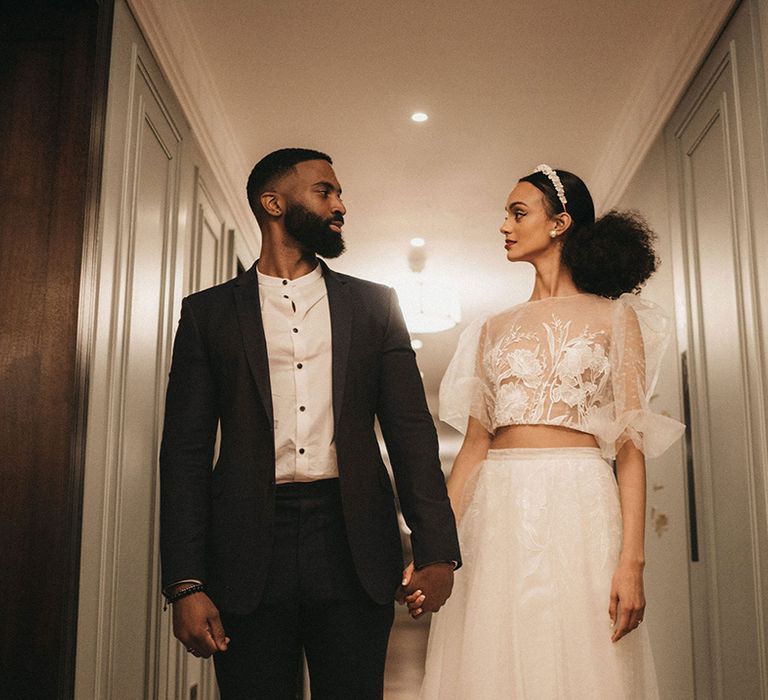 Black groom in an open collar shirt and forest green suit holding hands with his Black bride in lace bridal separates 