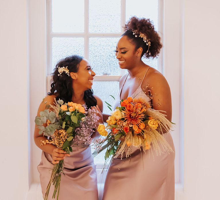 Bridesmaids in dusky rose satin bridesmaid dresses holding yellow and orange flower bouquets and wearing jewelled hair accessories 