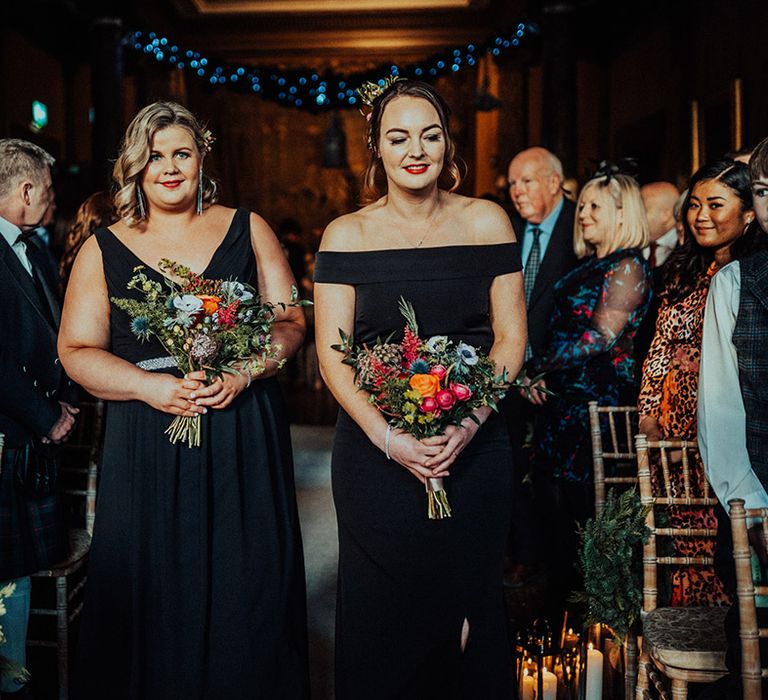 Bridesmaids in black bridesmaid dresses walk the aisle together holding bright pink, blue and orange flower wedding bouquets