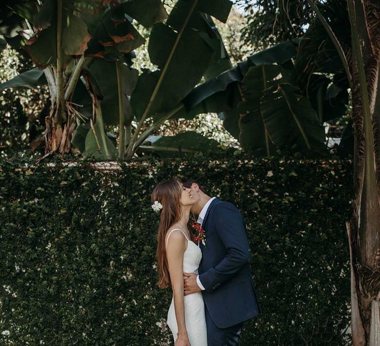 Bride in v-neck fitted wedding dress with fishtail train and bright red wedding bouquet with groom in blue suit and white trainers