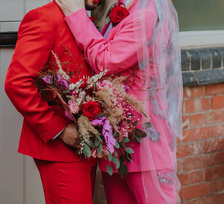 Stylish groom in a pink two-piece suit with bow tie, and embroidered wedding veil embracing his partner in a red suit holding a pink wedding bouquet 