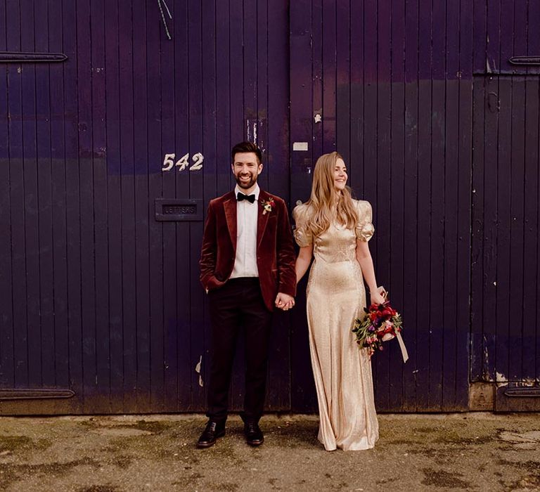 Bride in gold puff sleeve wedding dress with bow detail intertwines her fingers with the groom in red suit jacket 