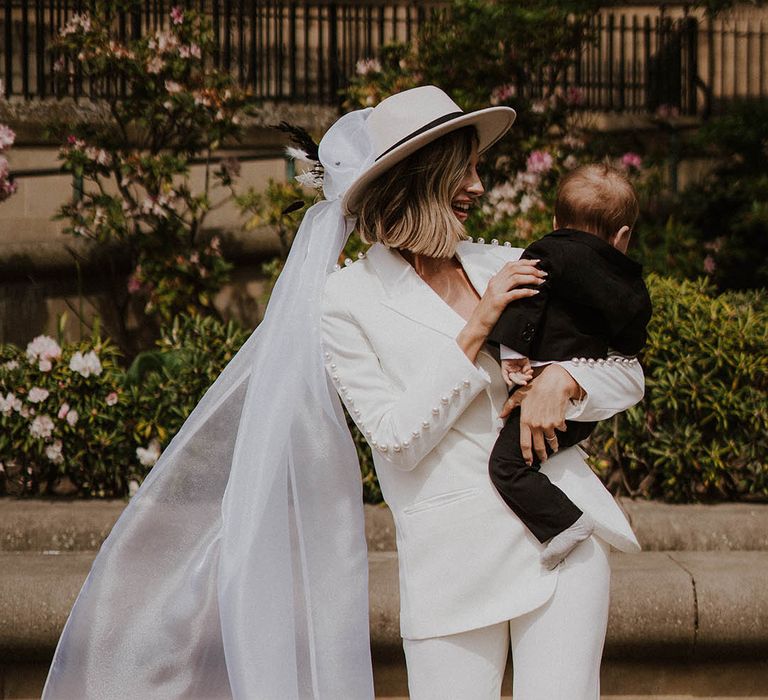 Bride with bobbed hair wearing white Nadine Merabi pearl trouser suit with veil attached holding son in black tie 