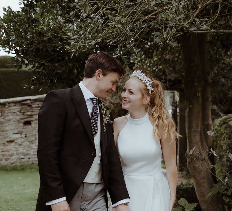 Bride and groom hold hands as they star at each other for their couple portraits with groom in morning suit and grey trousers and bride in a halter neck wedding dress 