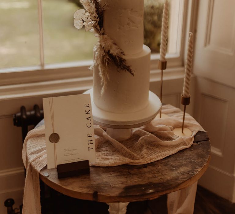 Two tier white wedding cake with dried grass decoration, twisted candles and a cake sign with a wax seal