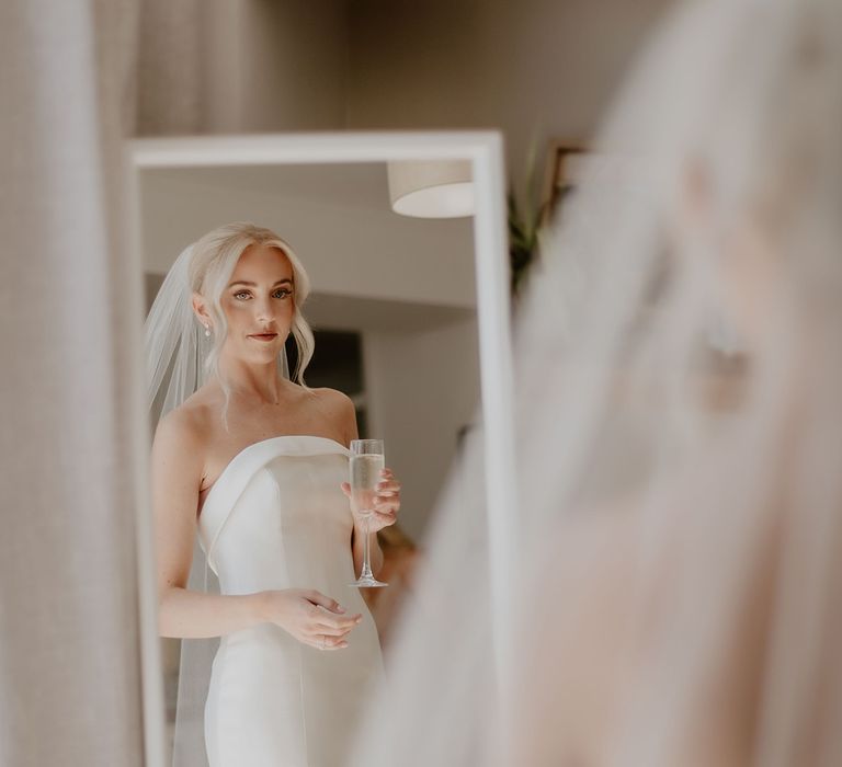 Bride in strapless wedding dress with pearl earrings and veil holds a glass of Prosecco as she looks in the mirror before getting married 