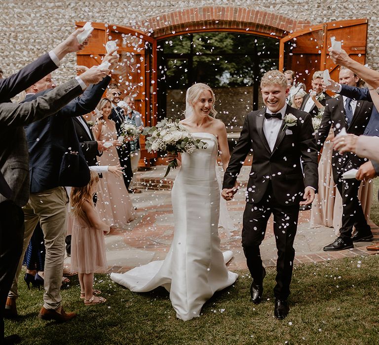 Bride in strapless wedding dress and groom in black tie smile widely as they have their confetti exit 