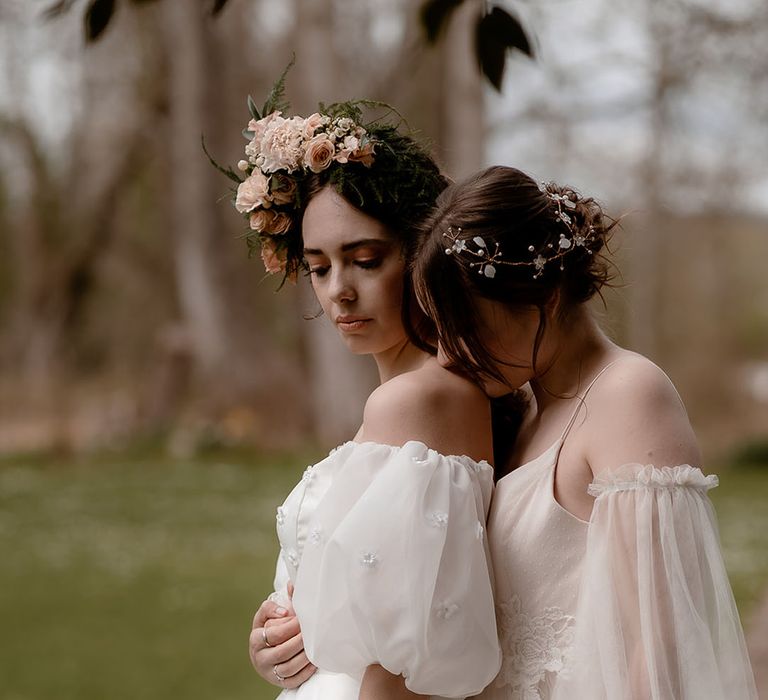 Bride in an ethereal wedding dress with sheer sleeves embracing her bride in a princess wedding dress with puff sleeves and flower crown