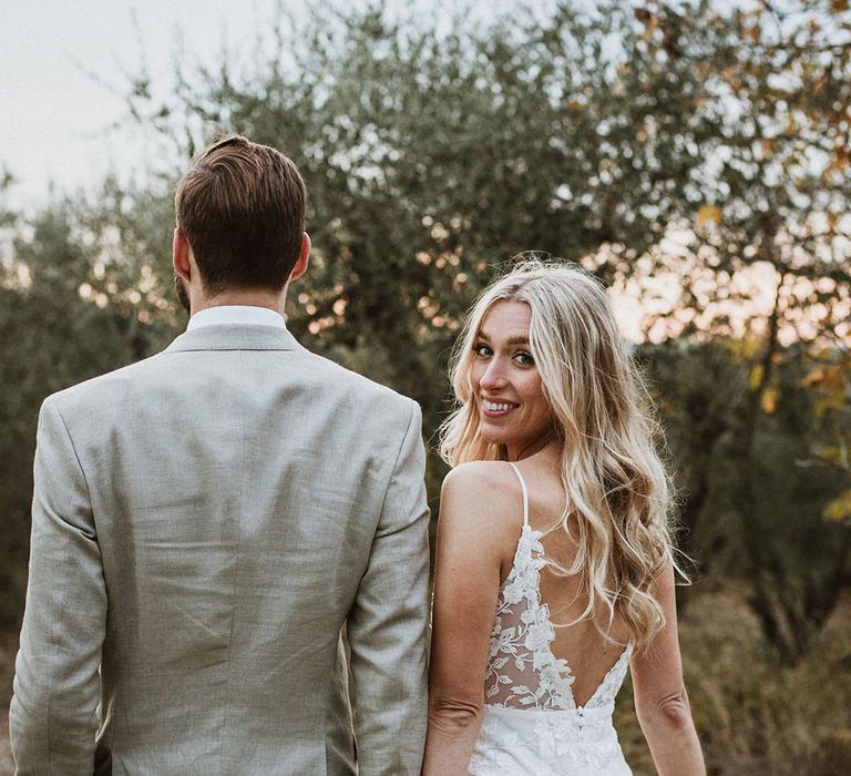 Bride holds train of wedding dress and looks back toward camera with her long blonde hair in loose curls 