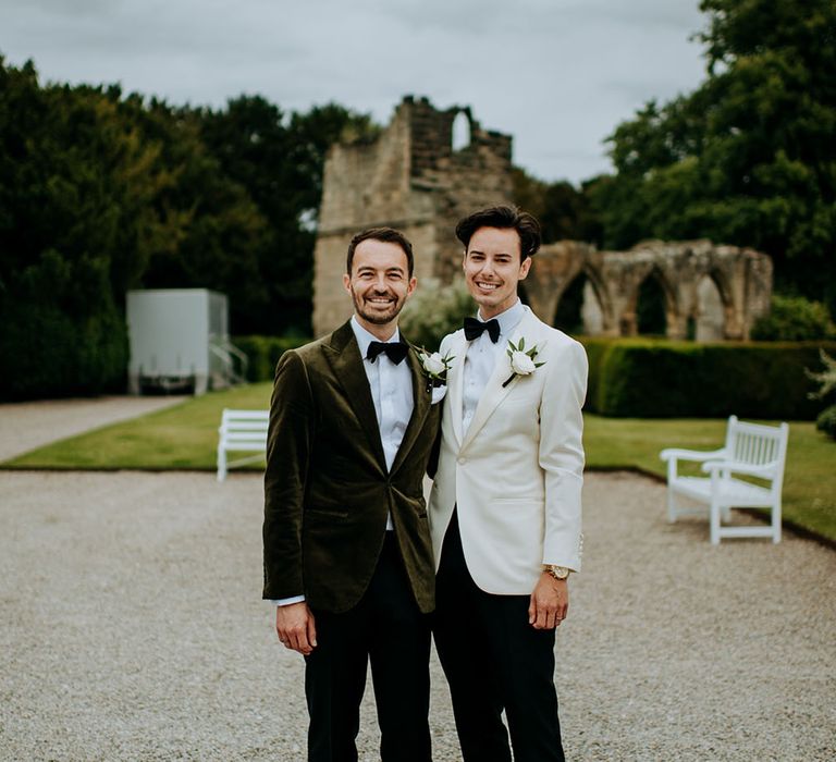 Groom wears green velvet dinner jacket and black tie stood beside his groom who wears white dinner jacket with their Jack Russell dog 