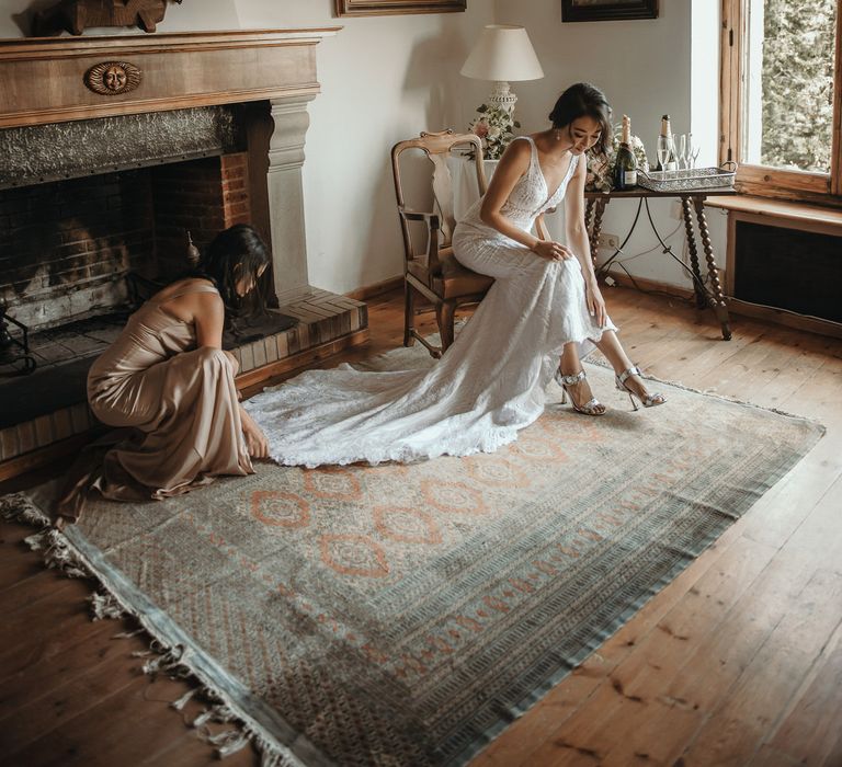 Bride puts on her Jimmy Choo silver peep toe heels on her wedding day as bridesmaid kneels beside her to fan out bridal train with scalloped edge 