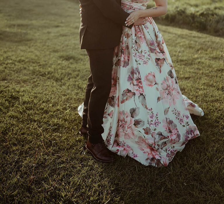 Bride wearing pearl hair pins and a floral wedding dress embraces the groom in a brown suit during golden hour 