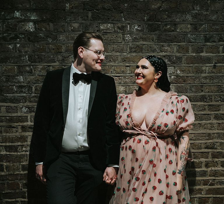 Groom wears velvet jacket and bow-tie as he stands beside his bride in pink chiffon dress with glitter strawberries 