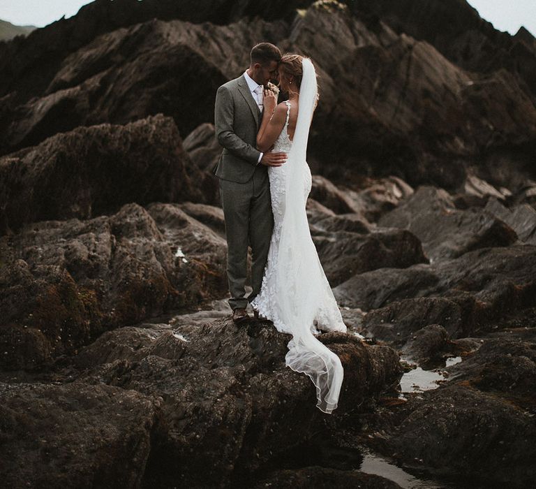 Bride wears low back fitted Enzoani wedding dress and floor-length veil as she embraces her groom on Tunnels Beach surrounded by rocks 