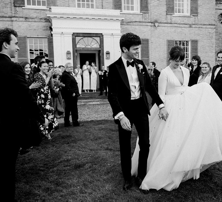 Groom in black tie and bride in princess tulle wedding dress walk through confetti after wedding ceremony at Findon Place 