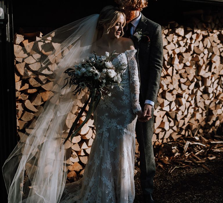 Bride holding a white flower bouquet in a lace mermaid wedding dress with the groom in a grey suit 