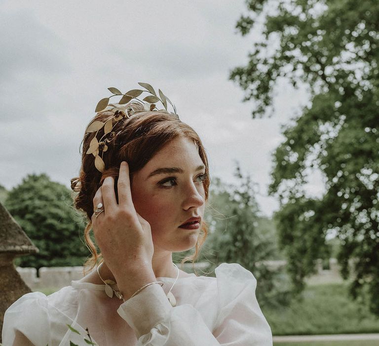 Bride wearing a sheer long sleeve wedding dress and gold leaf headpiece holding an oversized bridal bouquet with green foliage and white and yellow flowers 