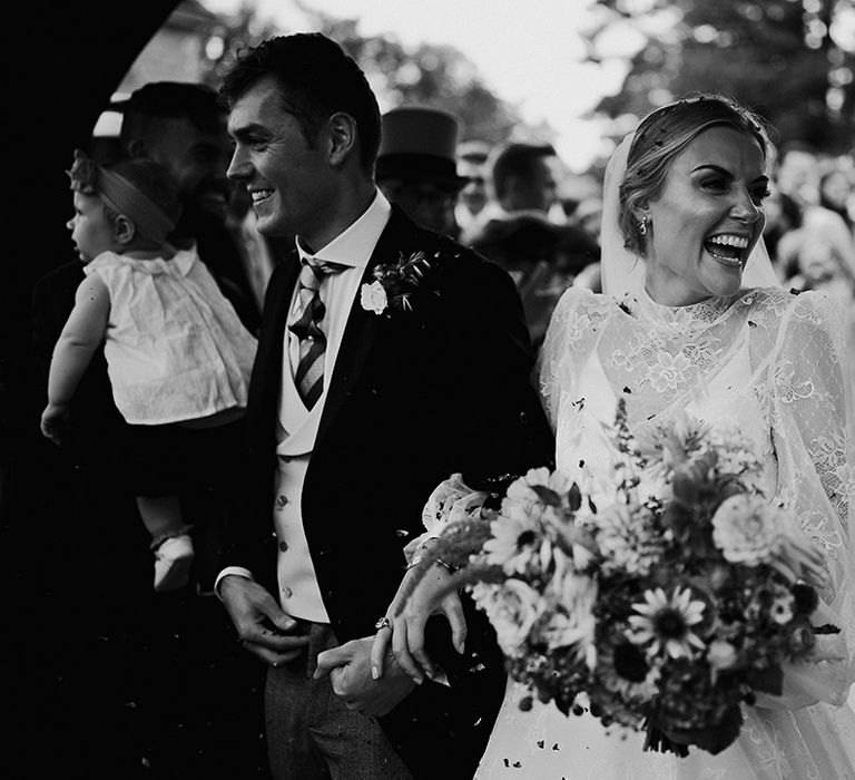 Groom in a morning suit and bride in lace wedding dress smile as they have their confetti exit 